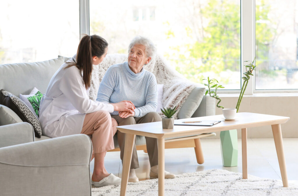 Pflegekraft berät Seniorin in ihrem Zuhause im Rahmen der ambulanten Pflege, während sie auf einem Sofa im Wohnzimmer sitzen.