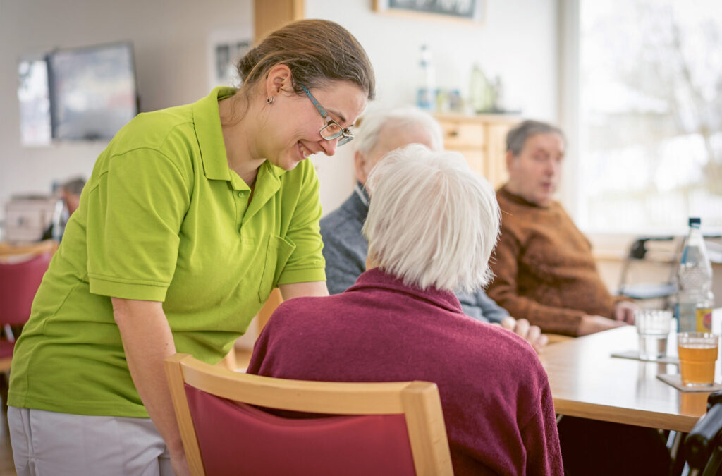 Pflegekraft betreut Seniorin in einer Tagespflegeeinrichtung, während andere Senioren gemeinsam am Tisch sitzen.