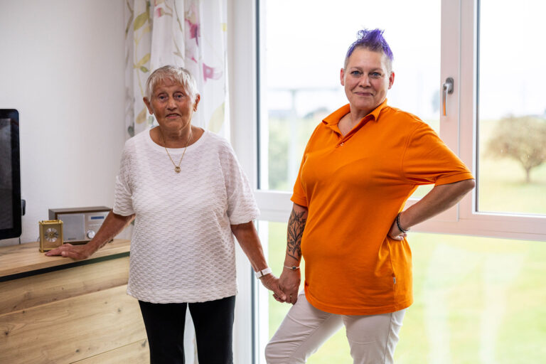 Pflegekraft in orangefarbener Uniform hält die Hand einer Seniorin und sorgt für Unterstützung und Vertrauen im Pflegeheim in Moosthenning.